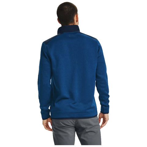 A person wearing a blue pullover is standing with their back facing the viewer while wearing gray pants in a neutral background setting.