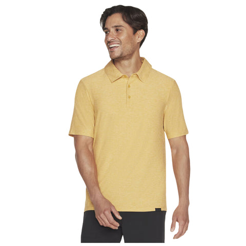 A smiling man wears a short-sleeved yellow polo shirt and stands with one hand raised slightly in a casual pose against a plain background.