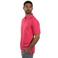 A man wears a bright pink polo shirt and light gray pants while standing sideways with one hand in his pocket and gazing slightly away in a neutral background.