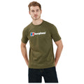 A man stands wearing an olive green t-shirt featuring the brand name "berghaus" in bold white letters with a blue and red logo on the left side while looking directly at the viewer.