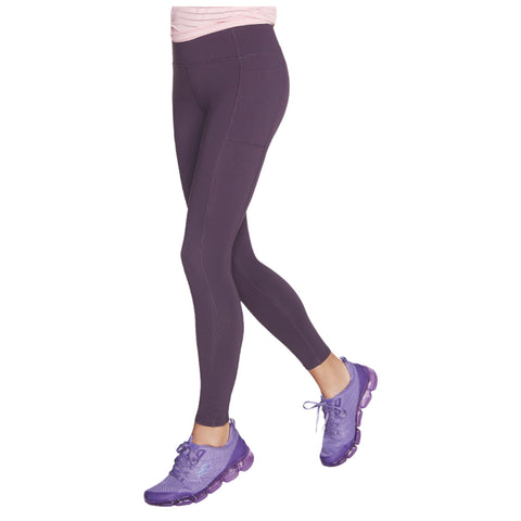 A person models dark purple leggings while walking. They wear matching purple athletic shoes and a light pink top, suggesting a casual exercise or active lifestyle setting.