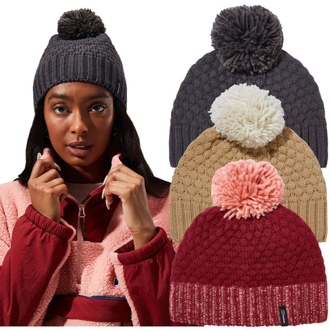 A woman wearing a pink and red jacket holds her collar while looking at the viewer. Four knitted beanies with pom poms are displayed behind her, showcasing different colors.