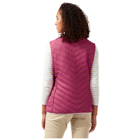 A woman stands facing away, wearing a pink quilted vest over a striped long-sleeve shirt, with light-colored pants in a plain background. Her curly hair is partially visible.