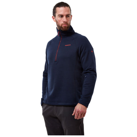 A male model stands wearing a navy blue pullover with a high collar and half zipper displaying a brand logo while looking to the side against a plain white background.