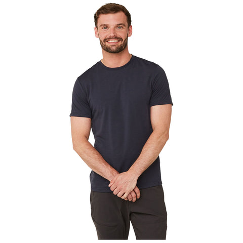 A man stands smiling with his arms crossed in front of him wearing a dark t-shirt and casual pants against a plain white background.