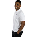 A man wearing a white polo shirt stands smiling with his hands in his pockets in a neutral background, showcasing a casual and confident demeanor.