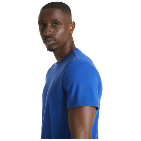 A man wearing a blue t-shirt stands facing slightly to the side with a serious expression against a plain white background. His hair is short and neatly styled.