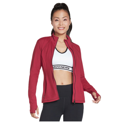 A woman stands smiling, wearing a red jacket over a white sports bra and black leggings, with her hands positioned slightly outward in a casual pose against a plain background.