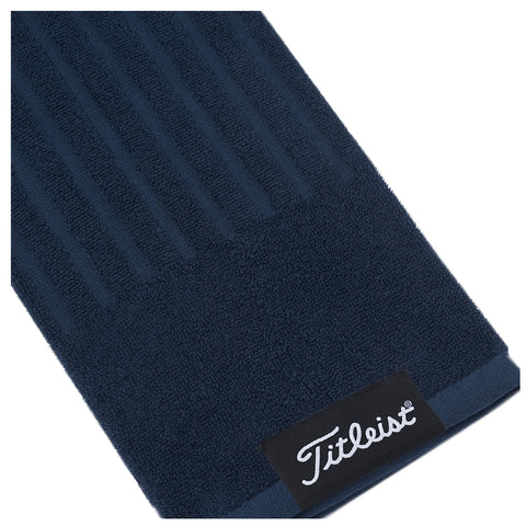 A dark blue towel with a textured pattern is positioned at an angle displaying the Titleist logo on a black tag indicating the brand identity and quality associated with golf.