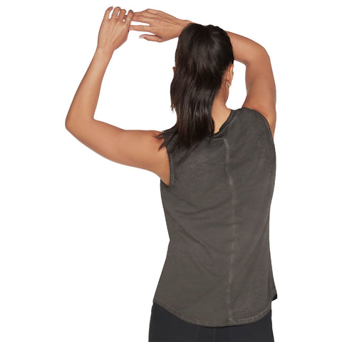 A person with long dark hair is stretching their arms overhead while wearing a sleeveless dark-colored top in a neutral environment with no visible distractions.