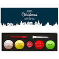 A boxed set contains colorful golf balls featuring festive designs and messages alongside white and red tees set against a dark background with a wintery village scene and holiday greetings.