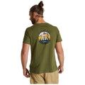 A person is wearing an olive green t-shirt featuring a design with mountains and trees on the back the text reads ADVENTURE GOOD FOR THE SOUL in a casual indoor setting.