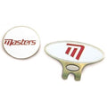 A round white golf ball marker displays the word "masters" in red text while an oval-shaped marker features a stylized red letter "M" on a white background both resting on a flat surface