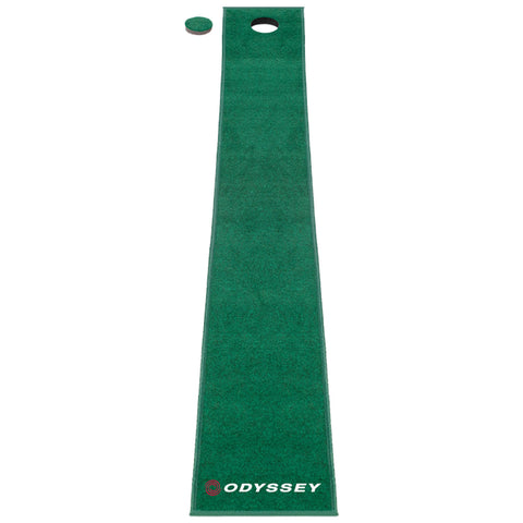 A long green putting mat lies flat with a hole at one end and a small golf ball beside it, set in an indoor environment for practice or play.