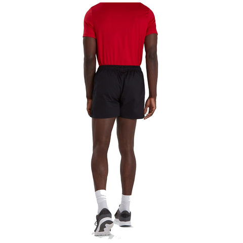 A person wearing a red shirt and black shorts stands facing away, showcasing athletic shoes and white socks in a plain background environment.