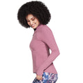 A woman with curly hair smiles while wearing a fitted pink long-sleeve top and patterned leggings, standing against a plain white background in a relaxed pose.