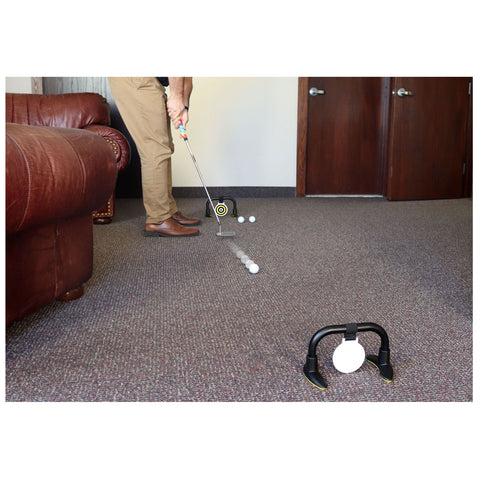 A golfer is putting a white ball with a club towards a target in a room with a brown couch and double doors in the background, surrounded by a textured carpet.