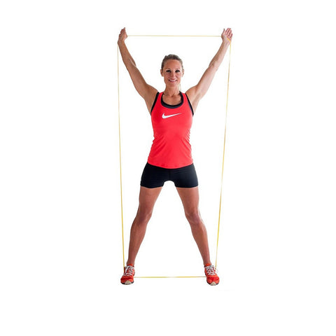 A woman in a red sports tank top and black shorts stands with a yellow resistance band held above her head, smiling against a plain white background.