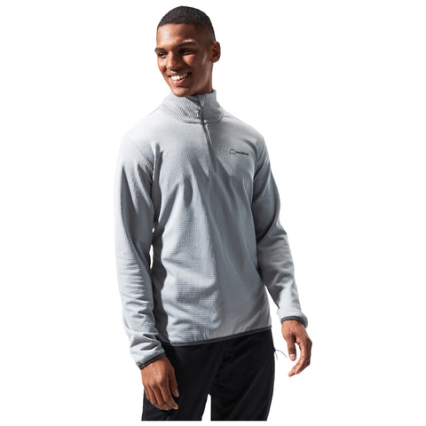 A young man wearing a light gray pullover with a zip collar stands smiling his arms relaxed at his sides against a plain white background.