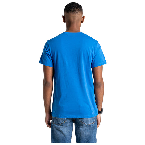 A person is standing with their back facing the viewer wearing a bright blue t-shirt and denim jeans in a neutral environment with a plain background.