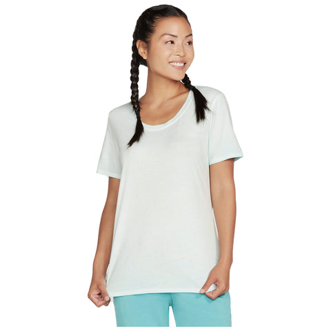 A woman with two braids smiles while wearing a light blue oversized t-shirt and matching pants, standing in a neutral background that emphasizes her relaxed and cheerful demeanor.