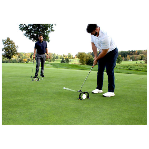 A golfer is putting a ball on the green using a training device while another golfer observes nearby on a well-maintained course surrounded by trees and hills in the background.