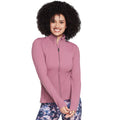 A woman with curly hair wears a pink zippered jacket and patterned leggings smiling as she poses with her arms crossed in a neutral background