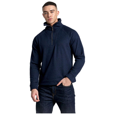 A young man wearing a dark navy quarter-zip pullover stands with one hand in his pocket and a serious expression on his face against a plain white background.