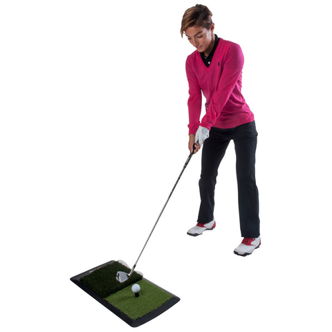 A person in a pink sweater swings a golf club striking a white golf ball positioned on a green turf mat in a practice setting.