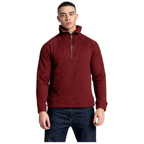 A man stands confidently wearing a dark red pullover with a half zip and a high collar paired with dark jeans in a plain white background.