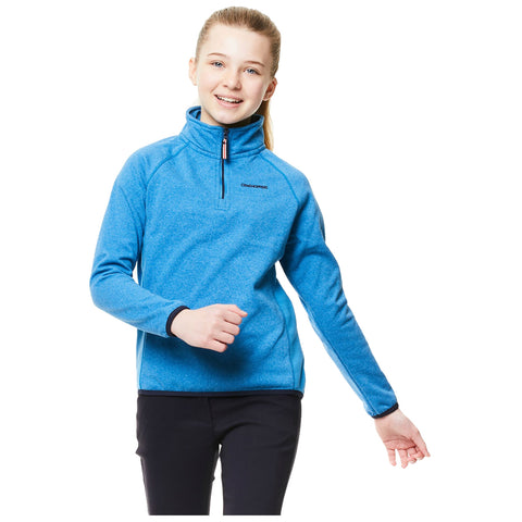 A girl wearing a bright blue pullover smiles while walking with one arm raised and a relaxed posture in a minimalist, brightly lit setting.