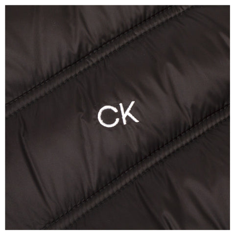 A black puffy jacket is displayed with a white embroidered CK logo prominently visible on the fabric's surface which features diagonal stitching patterns throughout.