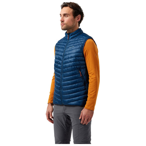 A man stands confidently wearing a blue puffer vest over an orange long-sleeve shirt and gray pants, conveying a casual yet stylish outdoor appearance.