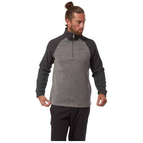 A man is adjusting the hem of his gray and black fleece pullover while standing in a neutral background. The pullover features a zippered neck and long sleeves.