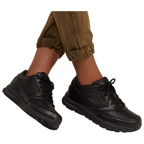 Black athletic shoes are positioned on a person's feet with brown pants rolled up at the cuffs revealing the shoes. The setting emphasizes casual footwear for everyday wear.