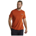 A man wears an orange t-shirt with a mountain graphic while standing with one hand in his pocket looking to the side in a neutral background