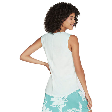 A woman with curly hair is smiling and looking to the side while wearing a light-colored sleeveless top and a patterned skirt in a bright and casual setting.