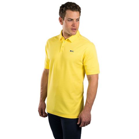 A man stands wearing a yellow polo shirt with a small logo on the left side, facing slightly to the right in a plain white background, looking confidently off to the side.