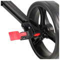 A black wheel is fitted with a red brake lever engaged to halt movement while metallic components and a frame are visible, suggesting it is part of a larger apparatus like a stroller.