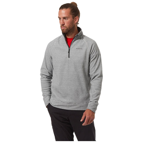 A man is standing confidently wearing a gray zip-up sweatshirt over a red shirt with a neutral expression his hands resting at his sides and black pants visible beneath the sweatshirt