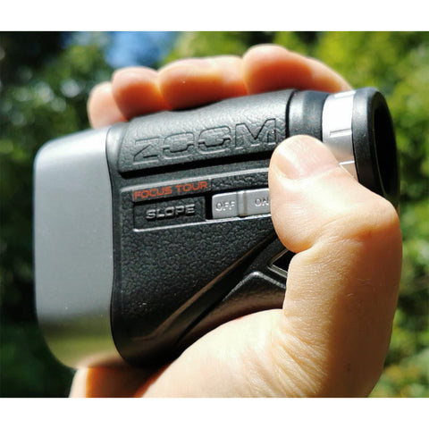 A hand is holding a handheld zoom device featuring buttons labeled SLOPE and FOCUS TOUR while surrounded by a lush green outdoor environment illuminated by sunlight.