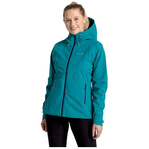 A woman stands smiling wearing a teal rain jacket with a hood and zippered pockets in a neutral background, suggesting a casual outdoor or active lifestyle.