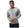 A man wearing a grey sweatshirt smiles and gestures with his hands against a plain white background showcasing casual attire and a friendly demeanor. The sweatshirt features a small logo on the chest.