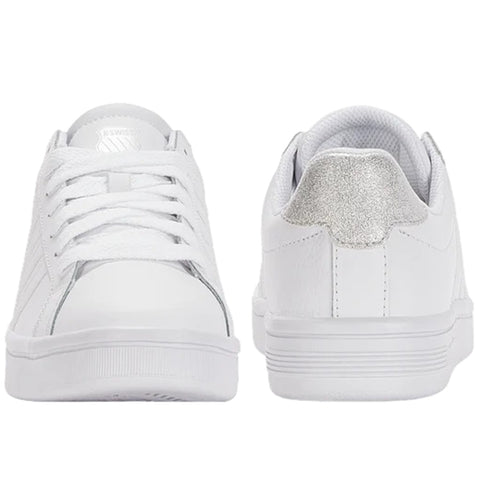 White sneakers are displayed with laces tied resting on a flat surface while showcasing a shiny silver accent on the heel contrasting against the sleek white design.