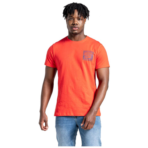 A young man stands confidently wearing an orange t-shirt with a graphic on the left chest and blue jeans in a simple bright background.