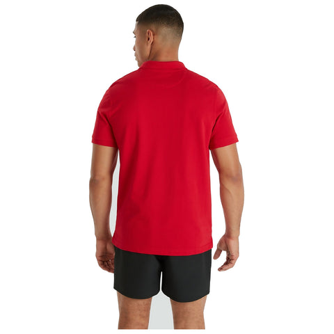 A man stands with his back to the viewer, wearing a red polo shirt and black shorts. He looks slightly to the side, indicating a casual stance in a neutral setting.