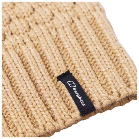 A knitted tan beanie rests on a surface displaying a textured stitch pattern with a small black label featuring the word berghaus prominently attached on one side.