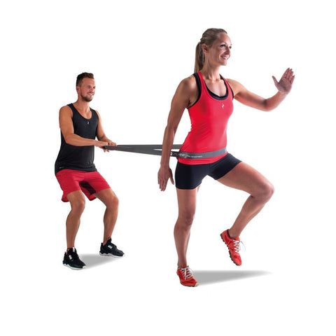 A man in a black tank top pulls a resistance band connected to a woman in a red tank top performing a high-knee exercise in a studio-like setting.