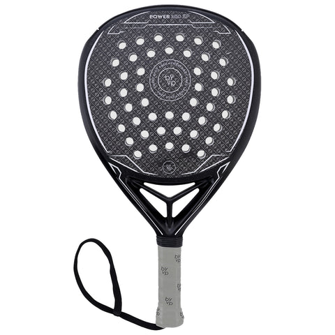 A padel racket with a large oval head featuring perforations for improved ball control is resting upright on a flat surface its grip wrapped in light-colored material with a wrist strap attached.