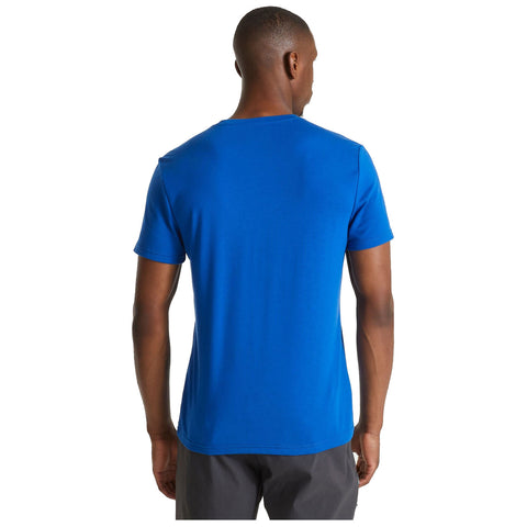 A blue t-shirt is displayed on a person seen from the back standing still in a neutral environment, showcasing a simple design and short sleeves.